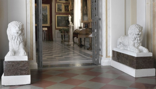 Widok na wnętrze pałacowe. Na pierwszym planie, po bokach drzwi wejściowych do drugiej sali - dwie rzeźby leżących lwów. W głębi sekretarzyk i galeria z obrazami.